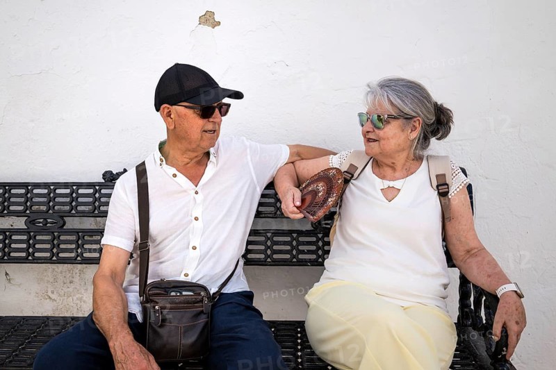 An active senior couple sits and chats on a bench in a picturesque white village in Spain. Dressed for comfort and travel, they enjoy a sunny day in an authentic Mediterranean setting