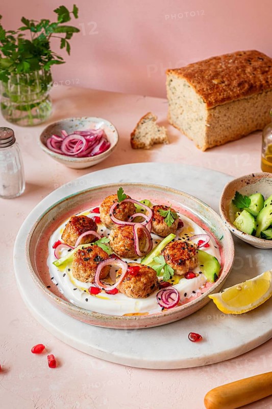 A beautifully arranged plate of meatballs garnished with sliced red onions, greens, and pomegranate seeds on a yogurt creamy base. Served with fresh bread and cucumber slices on a light background