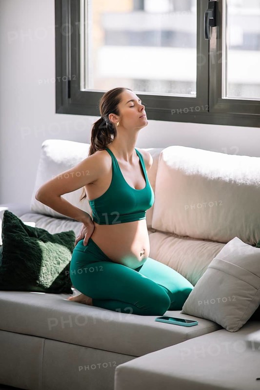 A pregnant woman in a green yoga outfit kneels on a sofa in a sunlit room, taking a moment to relax and focus on wellness. The setting emphasizes comfort and tranquility