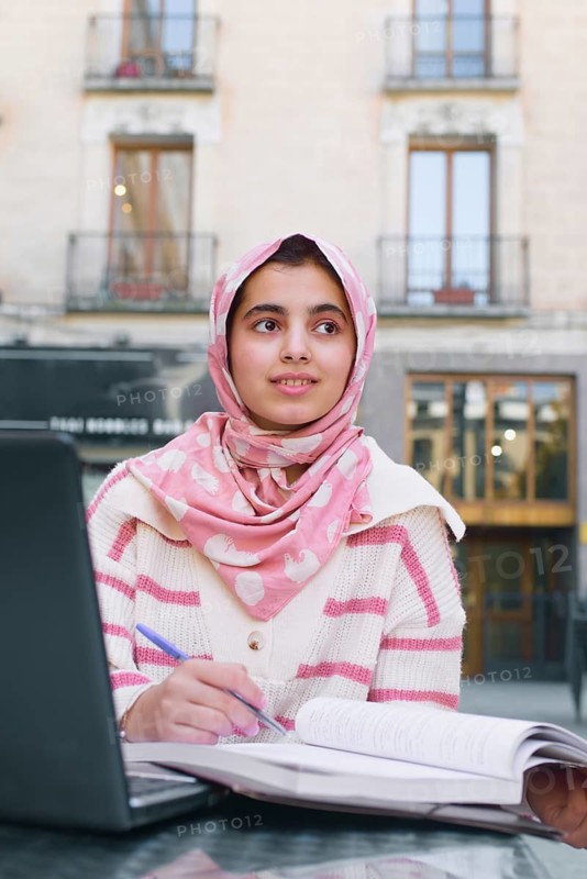 A arab student woman wearing a pink hijab studies with a laptop and open book outdoors. She holds a pen, appearing thoughtful. The setting is urban with blurred building in the background
