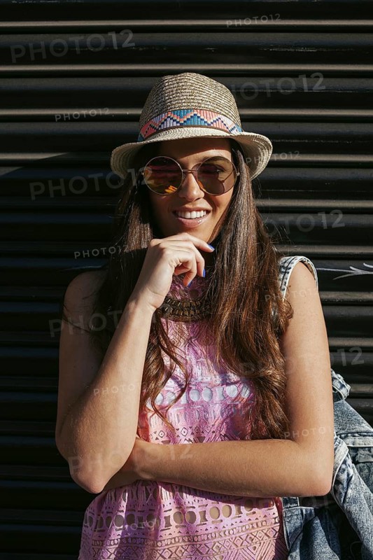 Fashionable woman in a straw hat and sunglasses, confidently posing against a dark, striped urban backdrop. Her relaxed summer style embodies carefree elegance