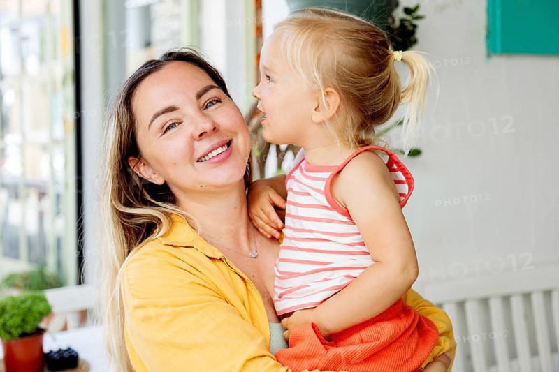 A smiling mother holds her young daughter in her arms while enjoying a sunny day outdoors. The child listens intently, showcasing a tender moment of connection and love