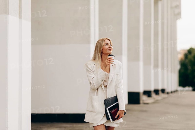 A businesswoman in a stylish white suit walks outdoors, holding a coffee cup, exuding confidence and a sense of purpose, against a backdrop of modern architecture