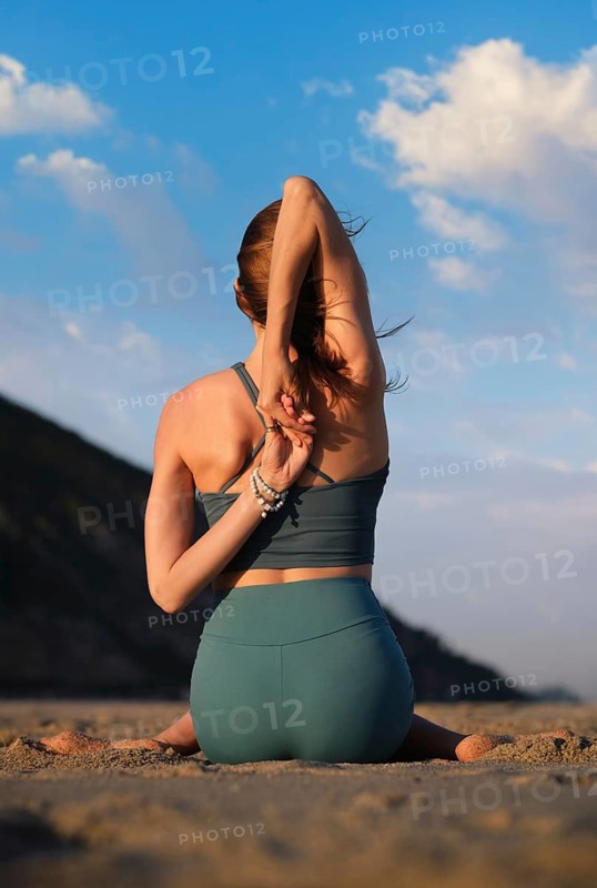 A woman gracefully sits in Gomukhasana, performing yoga by the beach during a stunning sunset, enjoying the tranquil view