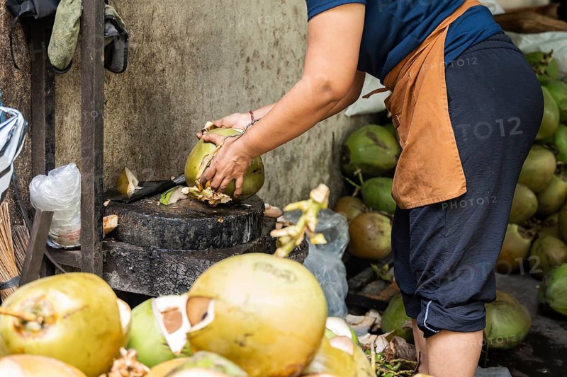 A vendor skillfully peels fresh coconuts at a street market using a chopping block. The scene showcases traditional preparation of coconut water, surrounded by ripe coconuts and tools