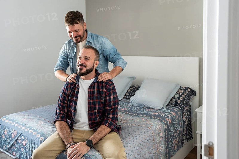 A joyful gay couple sharing a tender moment in their bedroom. One partner stands behind, gently massaging the other's shoulders, conveying love and companionship
