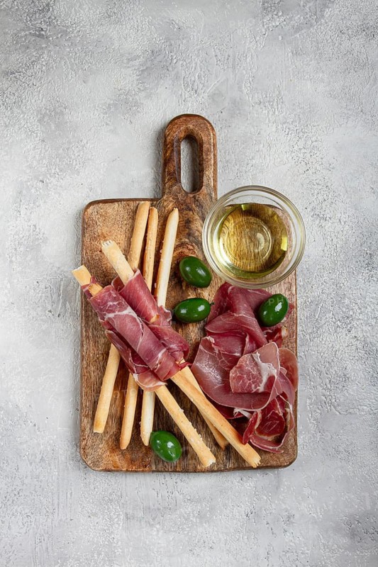 An appetizer of prosciutto, grissini and olives, served on a wooden board, with olive oil