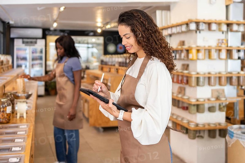 Two clerks in an organic supermarket efficiently manage inventory with digital tools. Shelves with jars and containers showcase a variety of bulk store products, enhancing customer experience