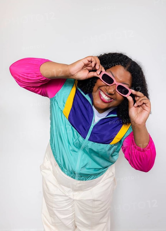 A joyful american woman wearing a vibrant windbreaker and pink sunglasses poses playfully against a white background, showcasing a retro fashion style with a big smile and positive energy