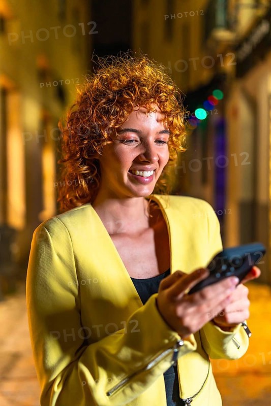 Vertical portrait of a happy woman with curly hair using phone in the city at night