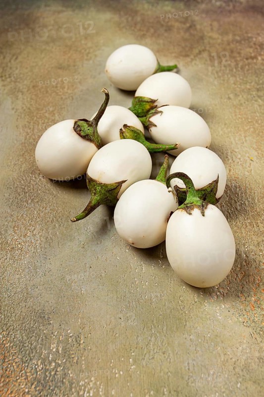 Several white eggplants are placed on a textured surface. The scene is well-lit and emphasizes the natural colors and shapes of each vegetable