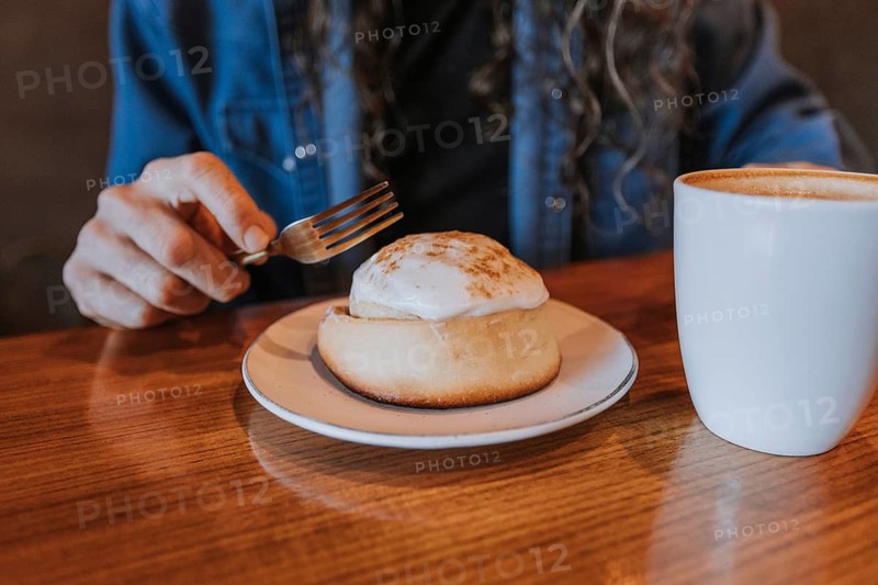 A man enjoys a cinnamon roll topped with icing alongside a steaming cup of coffee on a wooden table. The table creates a warm, inviting breakfast atmosphere
