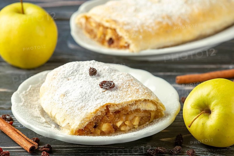 Apple strudel on plate with powdered sugar and raisins, next to yellow apples and cinnamon sticks
