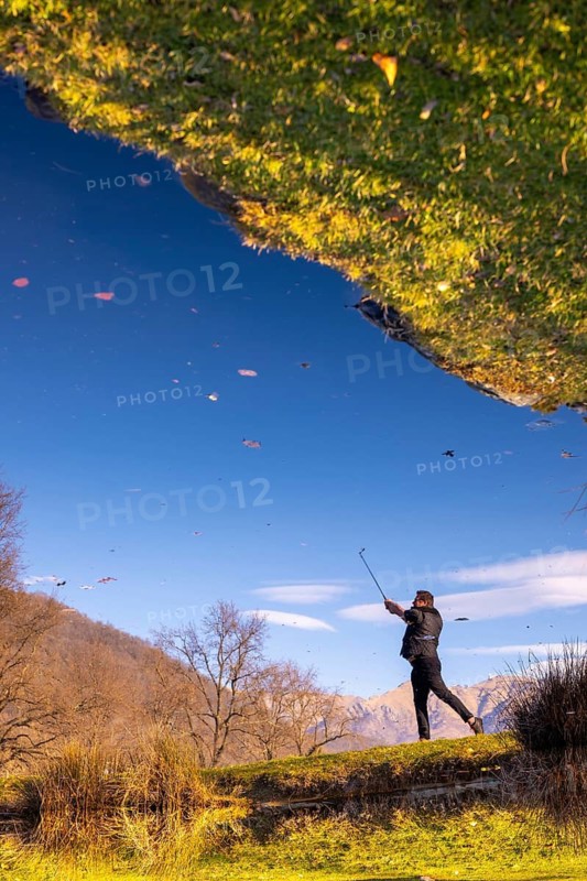 Male Golfer Reflected in a Water Pond and Hitting the Golf Ball on Fairway on Golf Course with Mountain in a Sunny Day in Switzerland