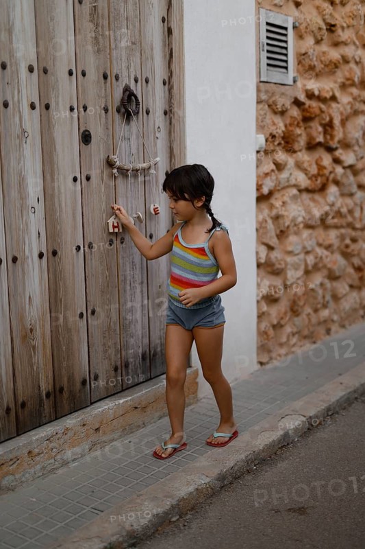 A young girl enjoys a playful moment at a wooden door in a quaint, rustic alley, engaging curiously with a door knocker