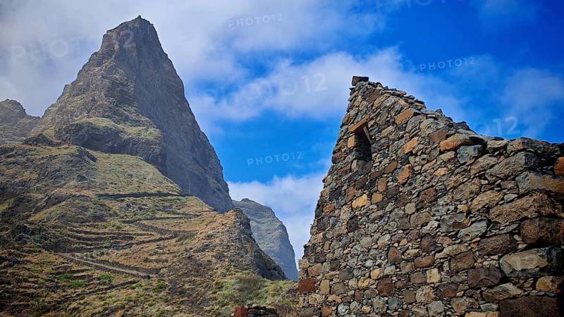 Stone ruin of a volcanic stone house in front of an impressive mountain, under blue sky, serpentine hiking trail in the background, hiking, lost place, abandoned village of Formiguinhas, Marinha da Cruzinha Natural Park, Santo Antao, Cabo Verde, Cape Verde, Cape Verde