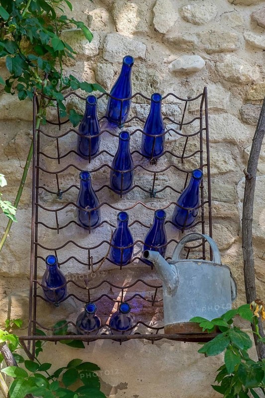 A wall shelf with blue bottles and an old watering can in front of a stone wall. Plants protrude into the picture from the side, Oppède-le-Vieux, Vaucluse, Provence Alpes Cote dAzur, France