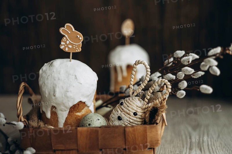 A traditional Easter setup featuring kulich, a Russian Easter bread, adorned with white icing and a bunny gingerbread cookie, alongside a basket filled with festive decorations including willow catkin branches and a handwoven straw rabbit