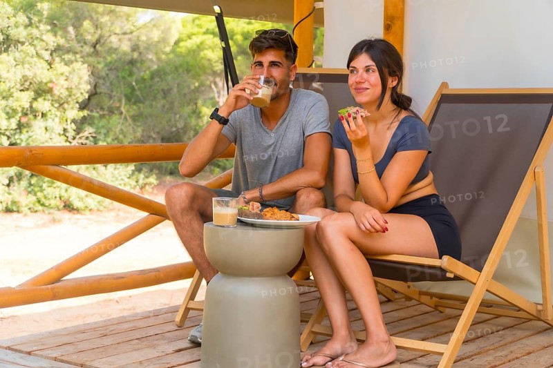 Young couple enjoying a leisurely breakfast on the terrace of their glamping tent, surrounded by lush greenery and the soothing sounds of nature, embracing a moment of tranquility and togetherness