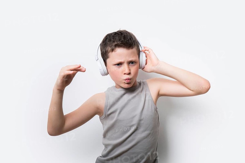 A boy in a gray tank top passionately listens to music on headphones, expressing joy and rhythm in his stance against a white background