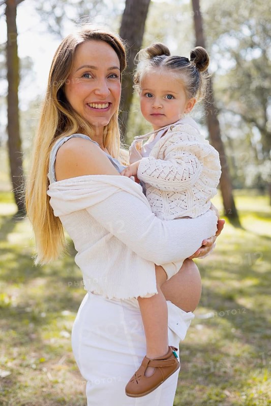 A pregnant woman holds her young daughter in a sunny outdoor setting. The mother and her child share a joyful moment amid the natural beauty of a park or forest