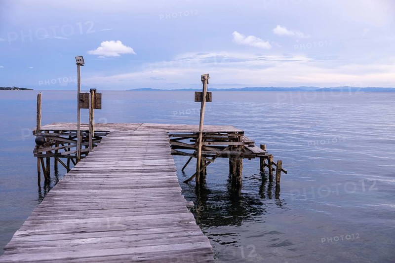 A serene wooden pier extends into the calm waters of Waigeo Island, Raja Ampat. The tranquil scene captures the island's natural beauty and peaceful ambiance