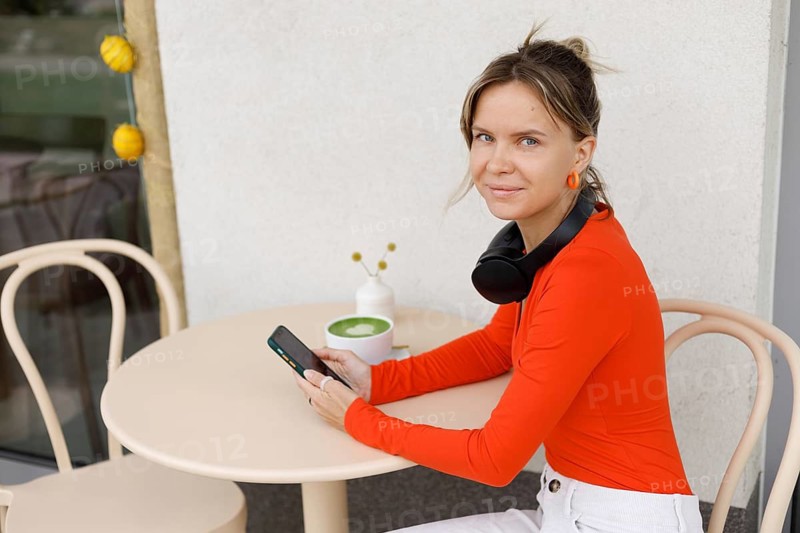 A woman in Warsaw sits at an outdoor cafe She is using her smartphone, with headphones around her neck and a latte on the table, enjoying a moment of relaxation