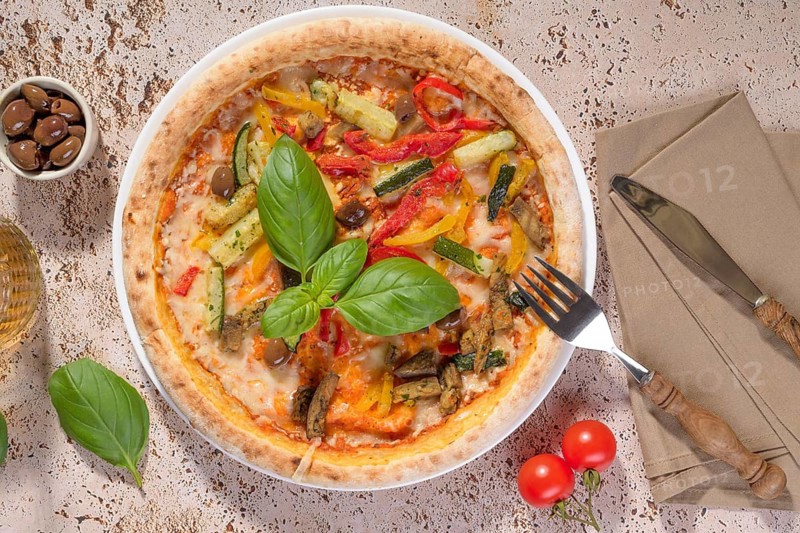 Mouthwatering vegetarian pizza topped with zucchini, eggplant, bell peppers, and olives. Fresh basil leaves garnish this Italian dish, served on a rustic plate with a side of olives