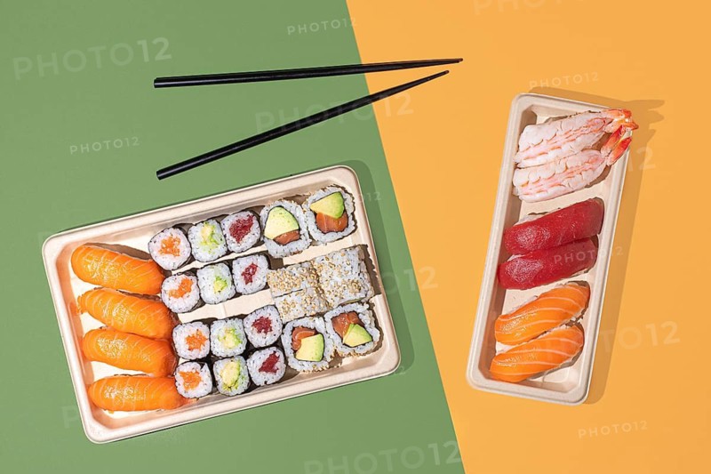 Minimalist overhead shot of sushi takeout boxes on a split color background, featuring nigiri and maki rolls with strong directional lighting