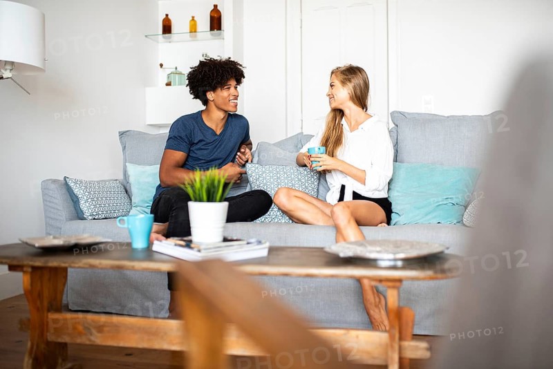A cozy living room scene with friends enjoying coffee and conversation. The room features modern decor, a wooden table, and decorative shelves. A relaxing moment indoors
