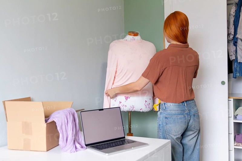 A woman prepares clothing for sale, using a laptop and dress form. A box nearby holds items for shipping, highlighting the process of online selling and closet organization