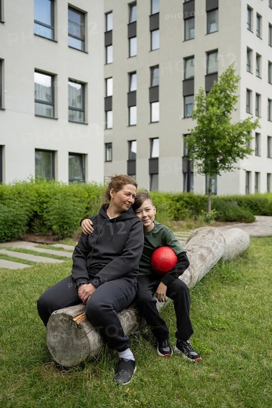 A mother and her young son sitting on a log outdoors, sharing a joyful moment. The boy holds a red ball as they enjoy the serene park setting, highlighting their bond
