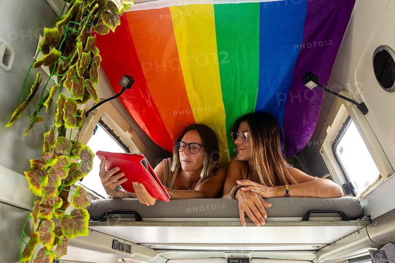 Two women relax in a camper van, using a tablet under a colorful rainbow flag. The vibrant scene captures leisure, technology, and diversity in a cozy setting