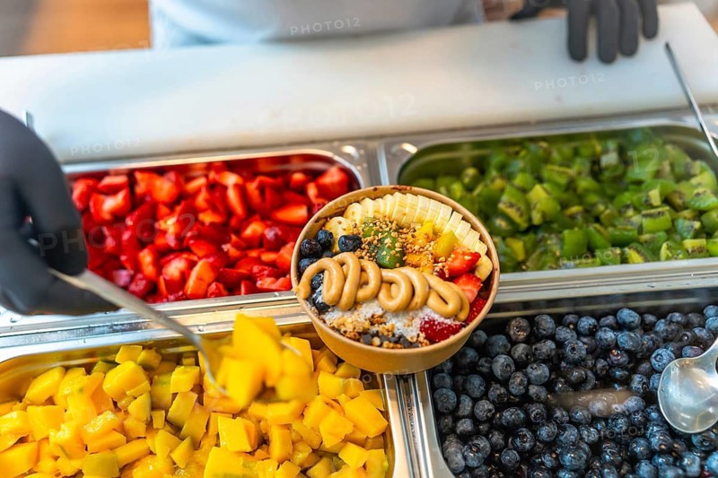 Chef in black gloves preparing an acai smoothie bowl, adding mango with a spoon from a buffet of fresh ingredients like strawberries, kiwi, blueberries, and bananas, topped with peanut butter