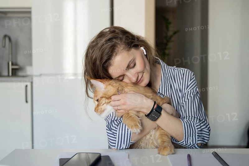 A woman embraces a ginger cat while sitting at a table in a cozy kitchen. She wears striped clothing and earbuds, exuding warmth and contentment in a relaxing home environment