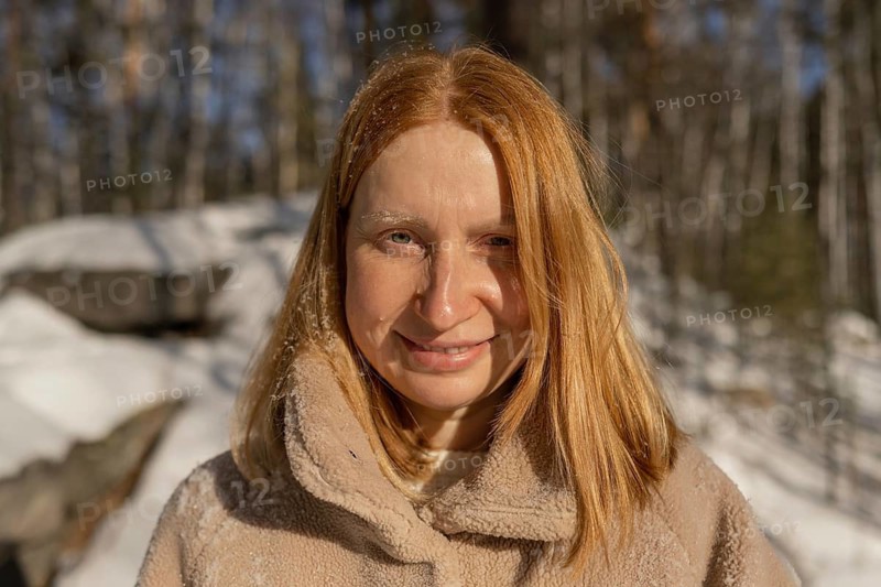 A smiling red hair woman looking at camera in a cozy jacket stands in a snowy forest, basking in the sunlight of a crisp winter day. The climate is evidently cold, yet her expression conveys warmth and contentment