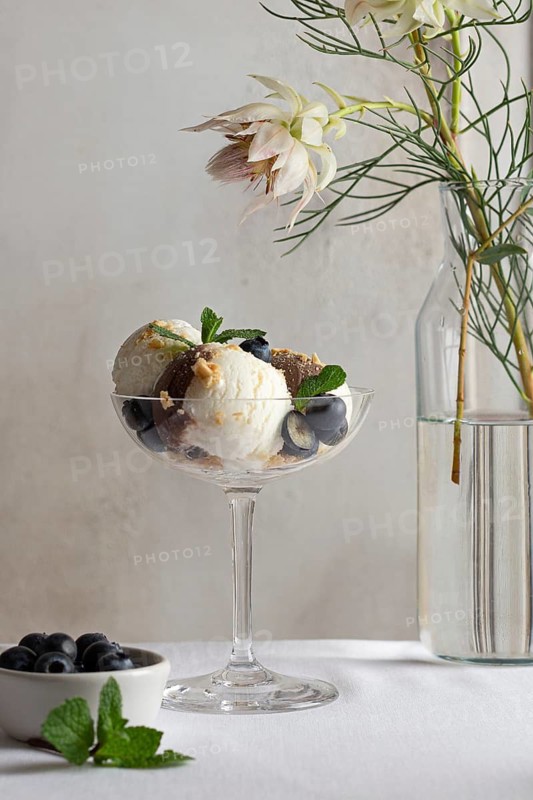 A sophisticated serving of vanilla and chocolate ice cream adorned with fresh blueberries, mint leaves, and chocolate pieces, presented in a stylish glass next to delicate white flowers