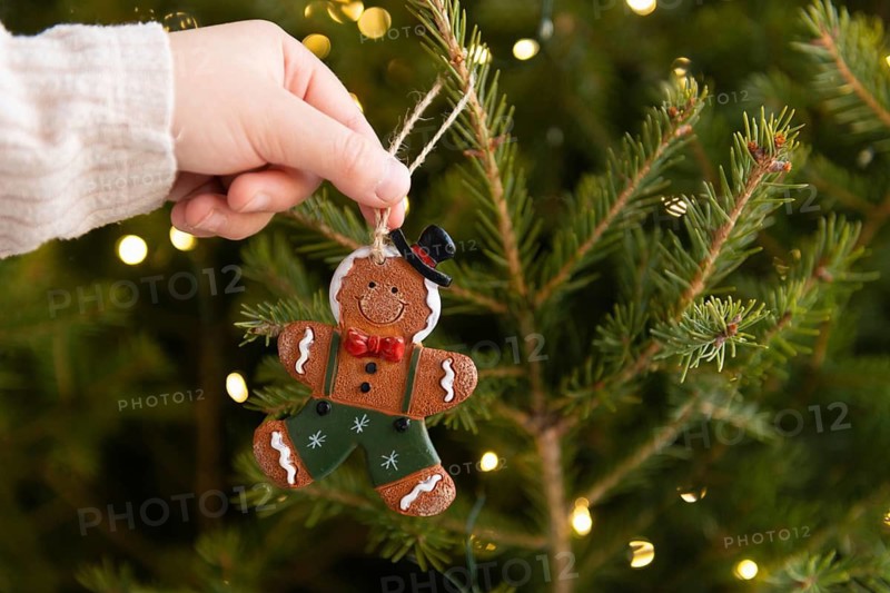 A close-up shot of a cropped unrecognizable child hand hanging a gingerbread man ornament on a beautifully lit Christmas tree, capturing the essence of family holiday celebrations