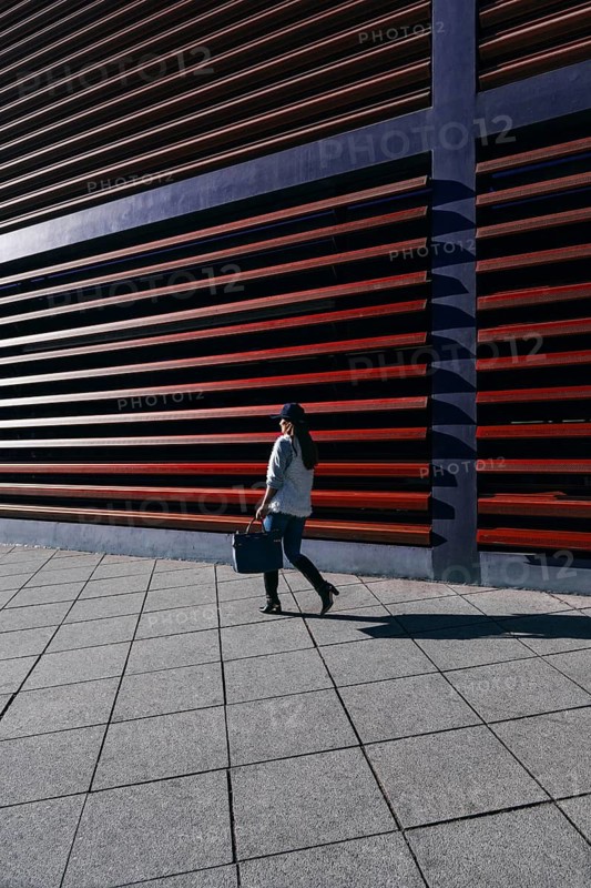 A woman in casual attire walks by a modern building with distinctive horizontal slats. The geometric alignment of the pavement enhances the architectural appeal