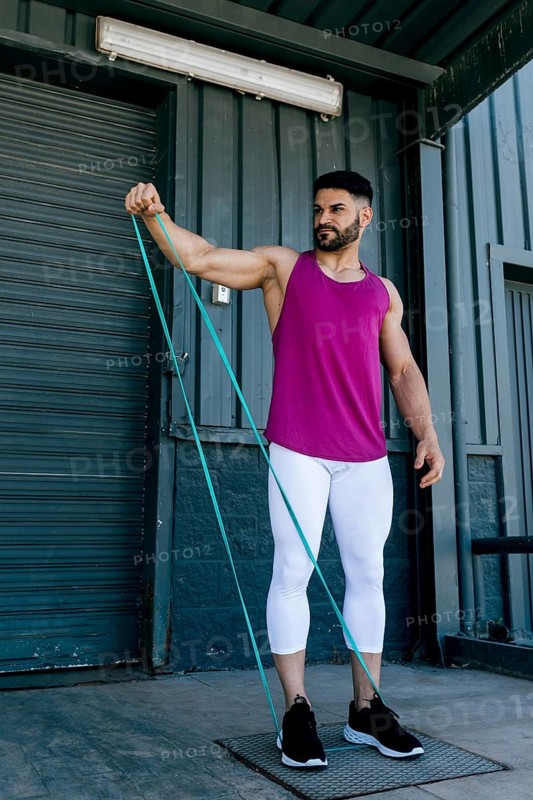 A fit man performs a shoulder exercise using a resistance band outdoors. Wearing athletic attire, he demonstrates strength and focus, promoting fitness and healthy lifestyle
