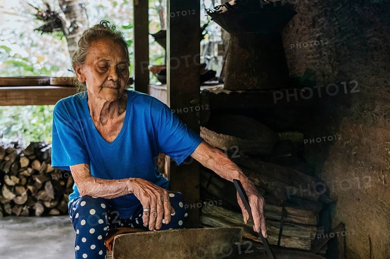 Elderly woman in a blue shirt sits next to a stack of wood and cooking balinese coffee, exuding warmth and wisdom. She seems focused and peaceful in her environment