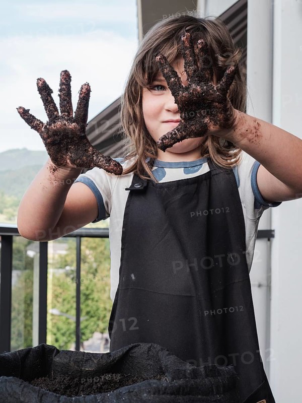 A young girl with messy soil covered hands proudly displays her dirty hands to the camera, symbolizing joy in gardening activities, with a soft-focus background of her garden environment