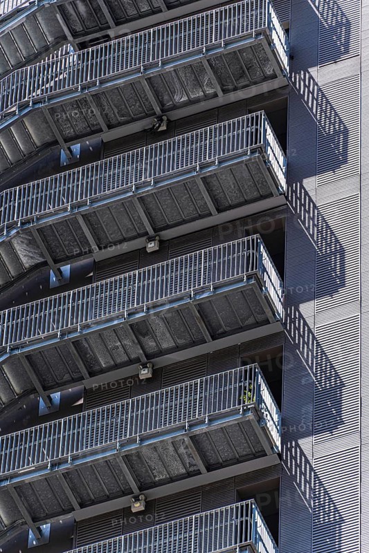 A series of sleek metal staircases adorn the exterior of an urban high-rise building, casting shadows on the modern facade. The industrial design exemplifies contemporary architecture