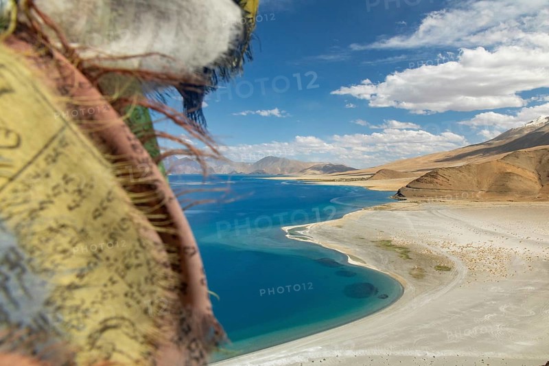 Breathtaking view of Pangong Tso lake in Ladakh, India, showcasing vibrant blue waters flanked by rugged mountains, under a bright sky, with prayer flags in the foreground
