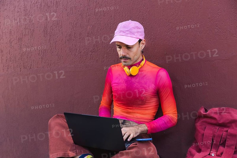 In Quito, Ecuador, a young Spanish man in pink clothing works on his laptop outdoors against a purple wall. His mustache and casual, colorful attire evoke an urban lifestyle with a touch of LGTBIQ+ flair, reflecting self-expression and diversity