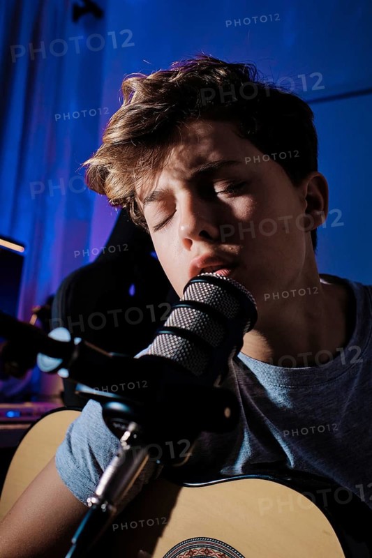A young musician, sings into a microphone while playing an acoustic guitar in home studio. The blue lighting and focused expression highlight their passion for music