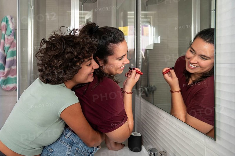 A loving lesbian couple shares a joyful moment in front of a bathroom mirror, with one applying lipstick and smiling while the other embraces lovingly