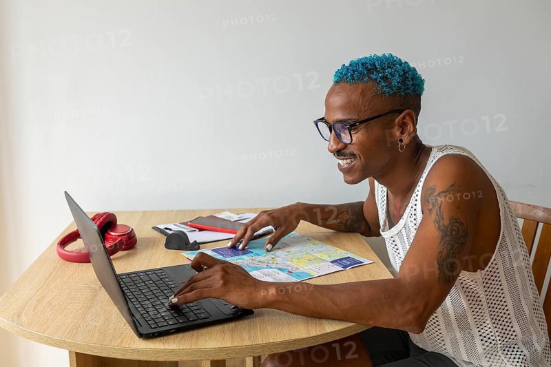 A smiling LGBTQIA+ individual with blue hair plans holidays using a laptop and map, embracing diversity and travel. The setting is casual and cheerful