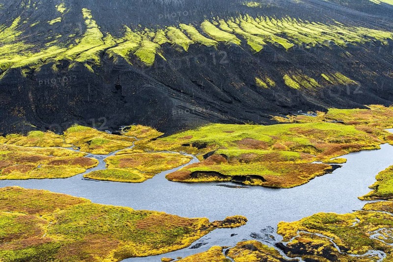 Aerial view of Iceland's riverside showcases unique textures with vibrant green moss contrasting against dark volcanic soil, highlighting nature's diverse and colorful beauty