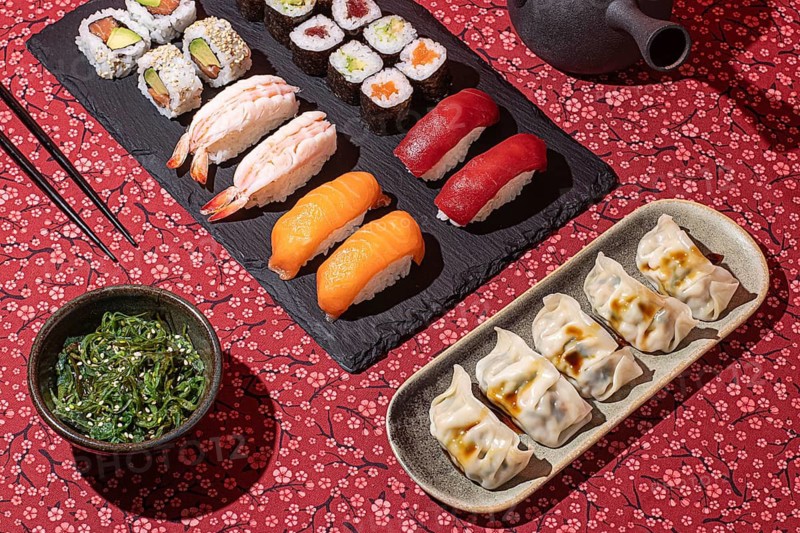 Flat lay of a traditional Japanese meal featuring nigiri, maki rolls and gyoza, presented on a floral red tablecloth with dramatic shadows
